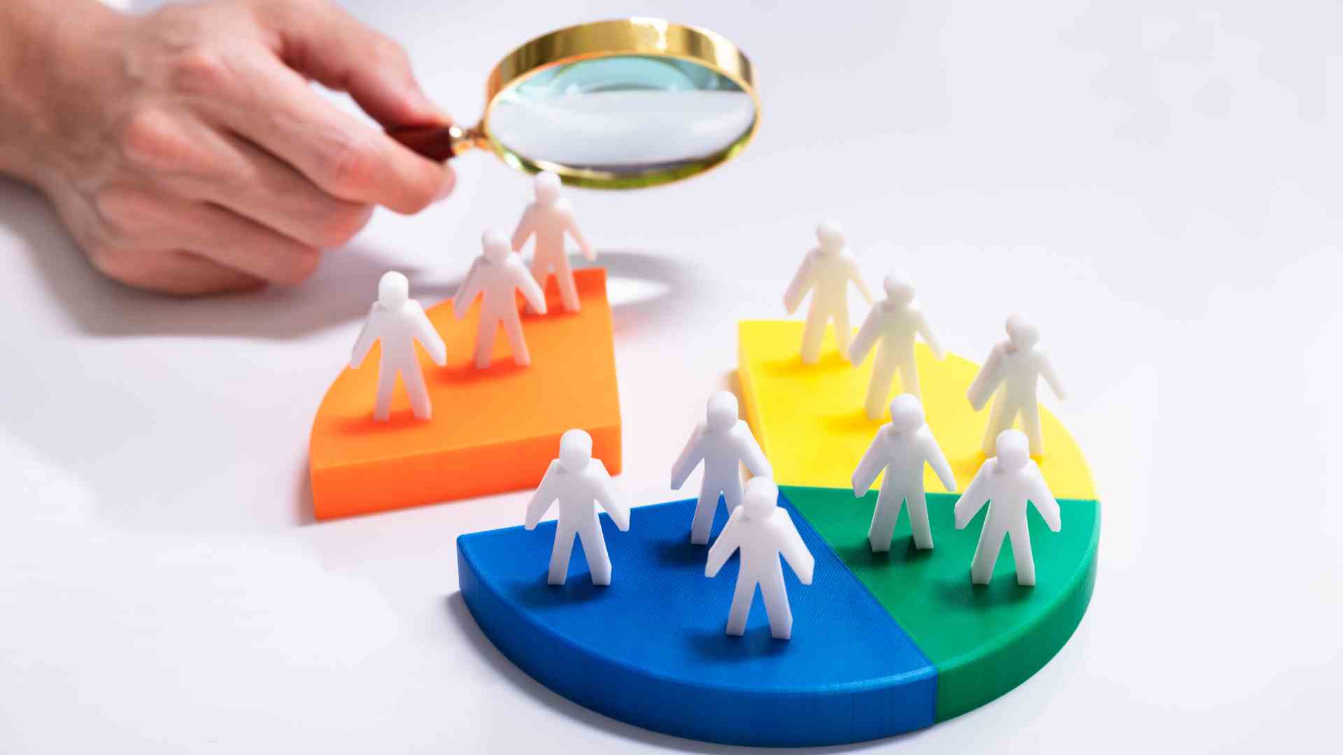 Magnifying glass examining groups of human figures placed on coloured pie chart segments, symbolising market segmentation and customer targeting.