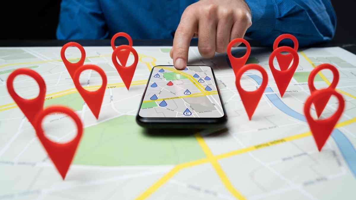 Photograph of a person’s hand pointing to a smartphone placed on a paper map. Around the phone are multiple red location pins, representing how people search for nearby businesses on maps. This highlights the importance of local SEO for small businesses.