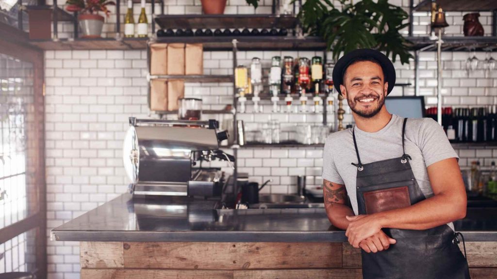 A café owner standing behind the counter in a modern coffee shop, smiling confidently. The image represents small businesses that rely on local SEO citations to help customers find them.